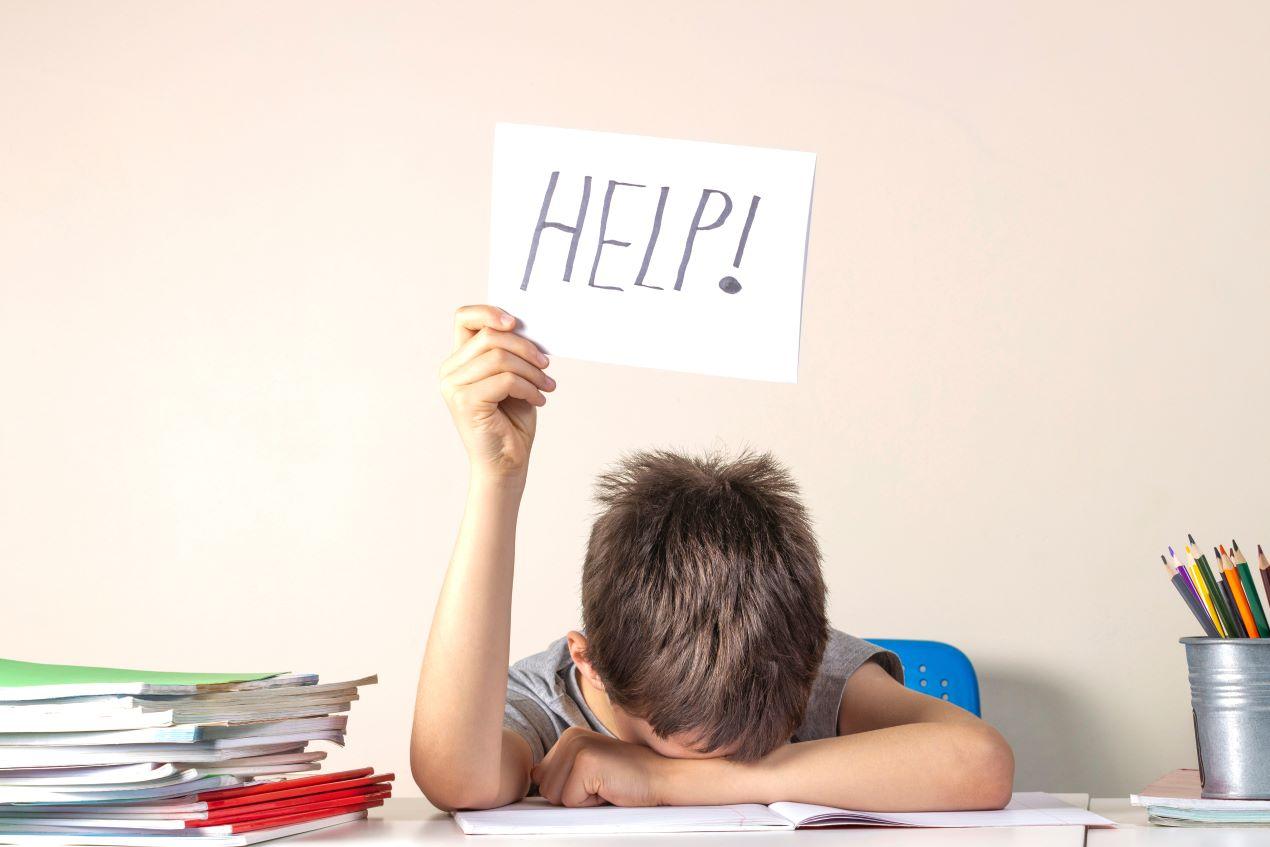 Ein Schüler hält erschöpft ein „HELP!“-Schild hoch, während er über seinen Schulbüchern liegt – Mathe Nachhilfe online kann die Lösung sein.