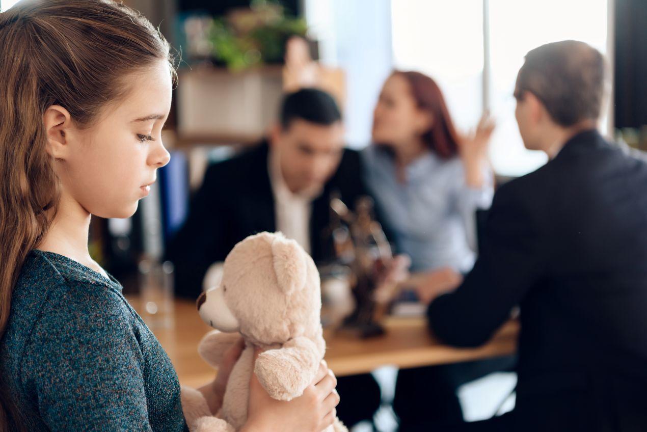 Ein Mädchen mit einem Stoffbären schaut traurig weg, während ihre Eltern vor einem Anwalt streiten – ein typischer Fall im Familienrecht Tauberbischofsheim.